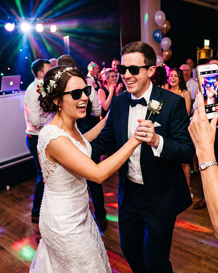 Bride and groom dancing in sunglasses while a guest photographs them on a phone