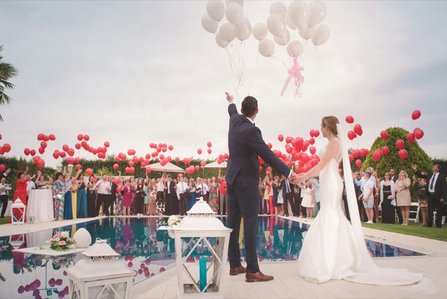 Newlyweds releasing red balloons by a pool with guests watching